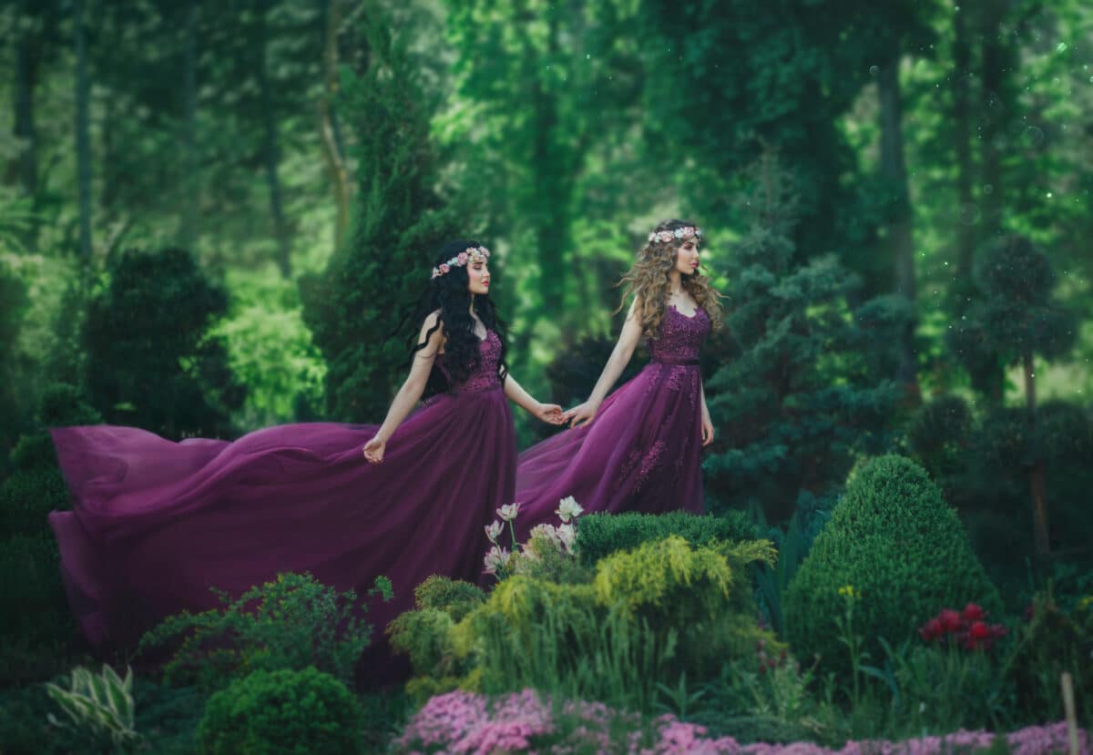 Two girlfriends, a blonde and a brunette, are holding hands. Background flowering garden. Princesses are dressed in luxurious purple dresses, with a long train, which are waving by a gust of wind