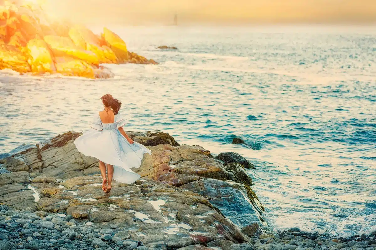 lady in white dress stands by the ocean at sunrise