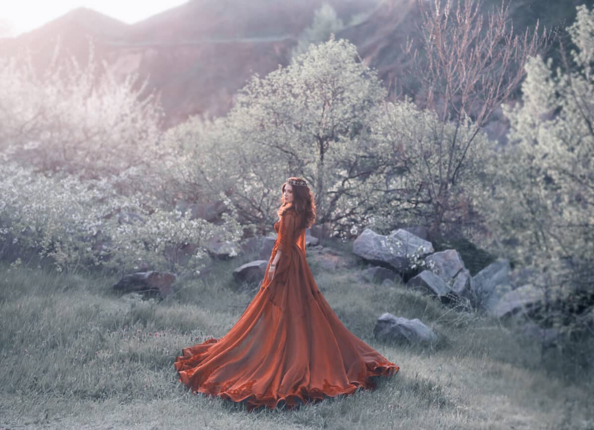 A brunette girl with a golden crown and in a red dress in a long train walking at sunset. Background wildlife, flowering trees, sunlight, lush grass