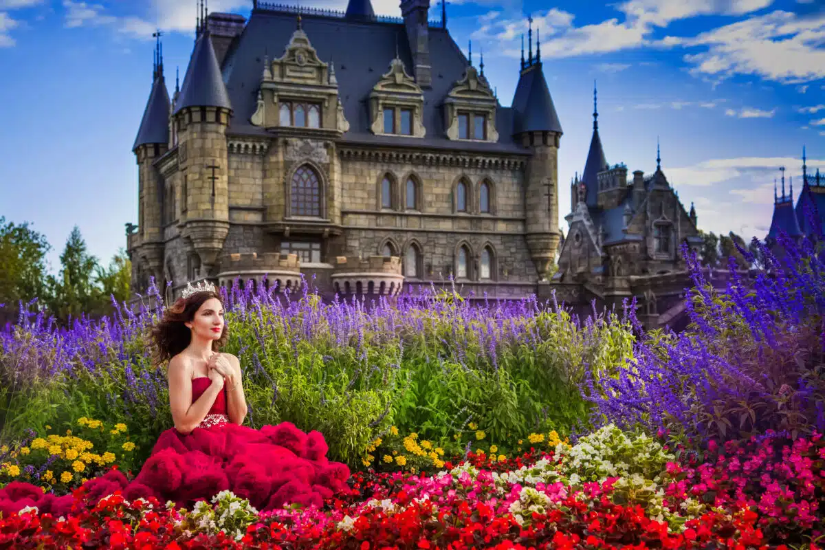 A beautiful woman, a princess in a red dress, sits by in a blooming garden. An ancient castle in the background. Medieval fantasy, European palace