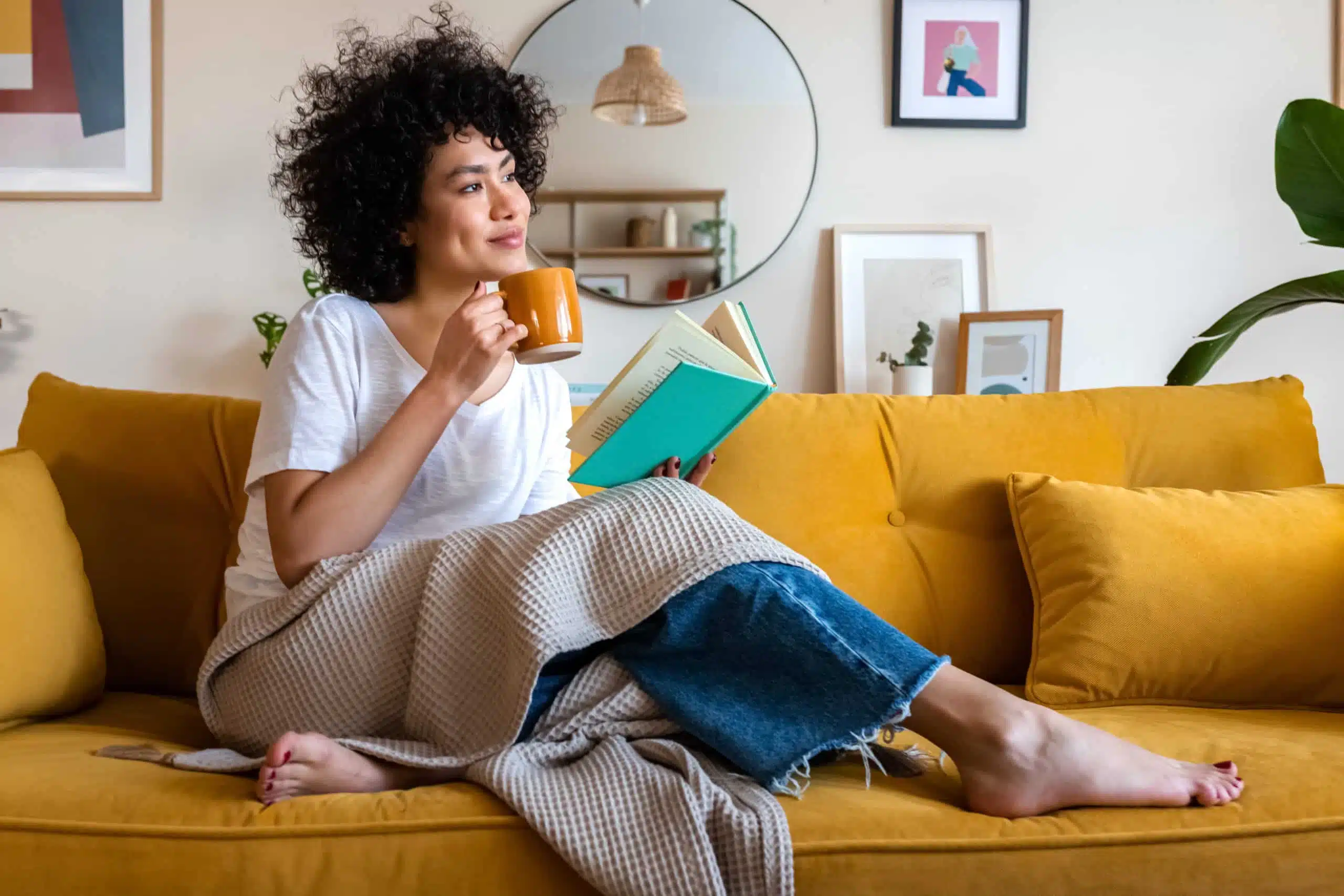 Pensive relaxed woman reading a book at home, drinking coffee sitting on the couch.
