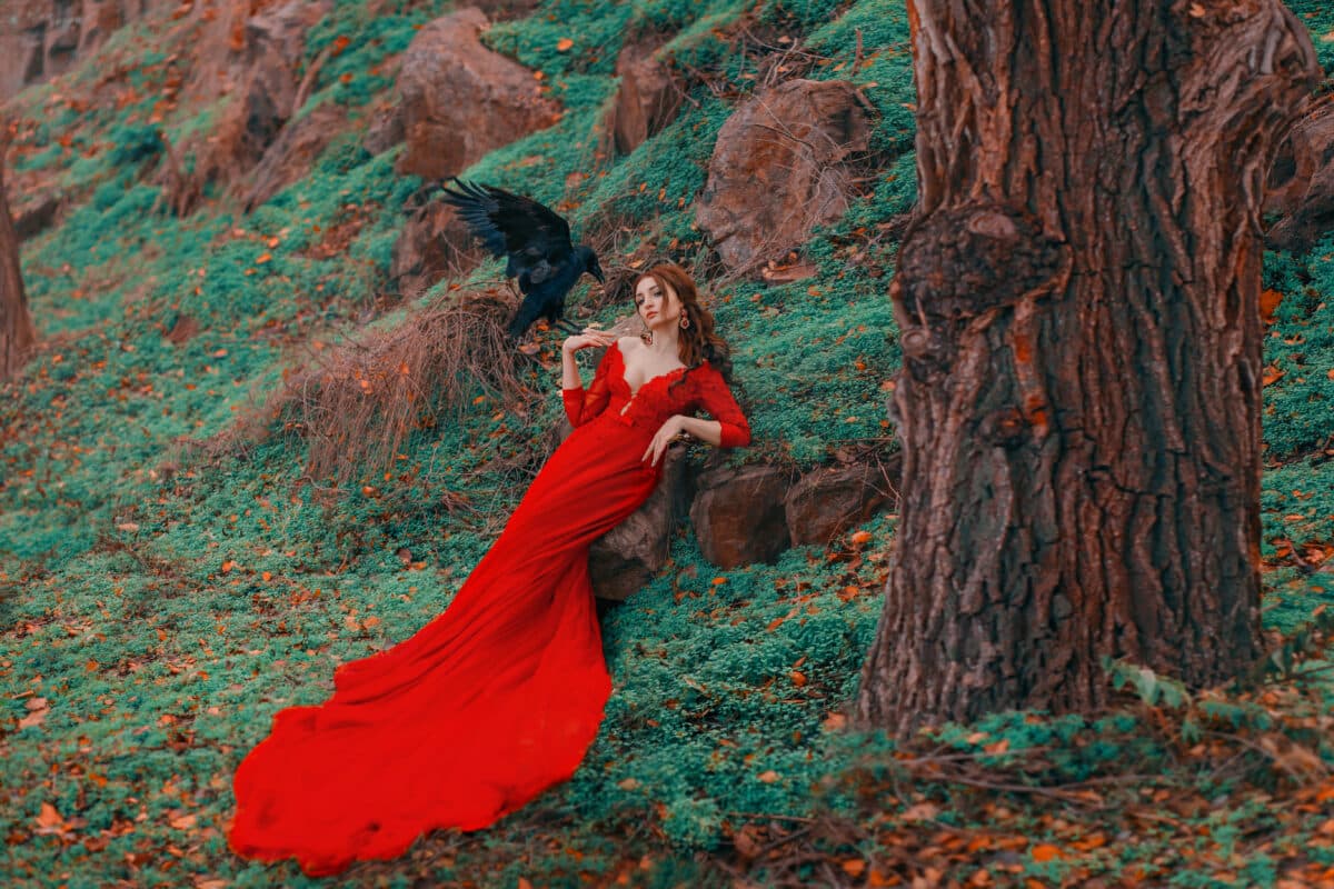 Beautiful sexy redhead woman sorceress in red long elegant dress with deep neckline is resting on green grass with fallen autumn orange leaves. Big Black Raven sits on magician’s hand and flaps wings.
