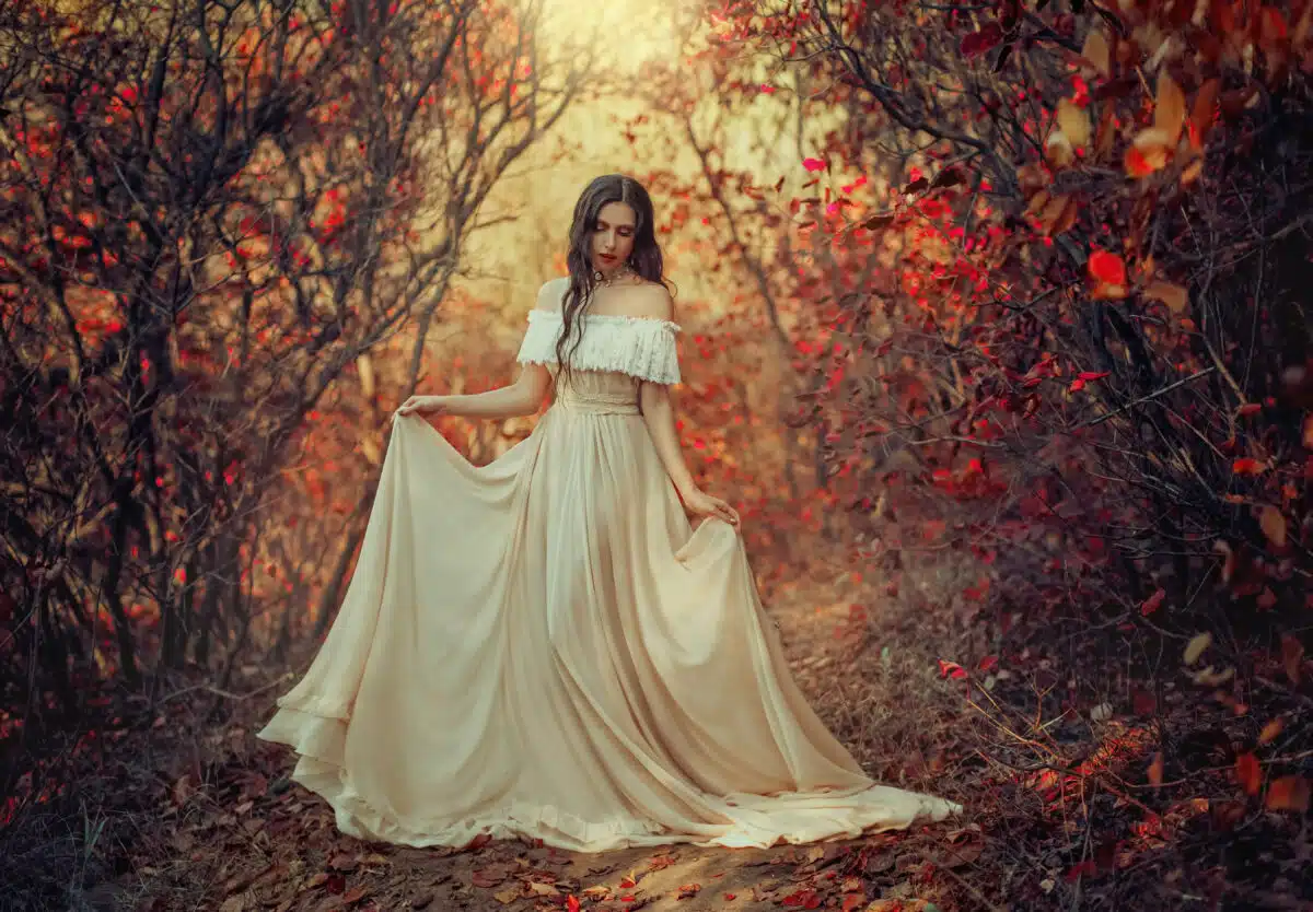 Art photo fantasy woman queen standing in gothic autumn forest, white vintage style dress. Girl princess beauty face long wavy hair, elegant sexy bare open shoulders. Red orange yellow color dark tree