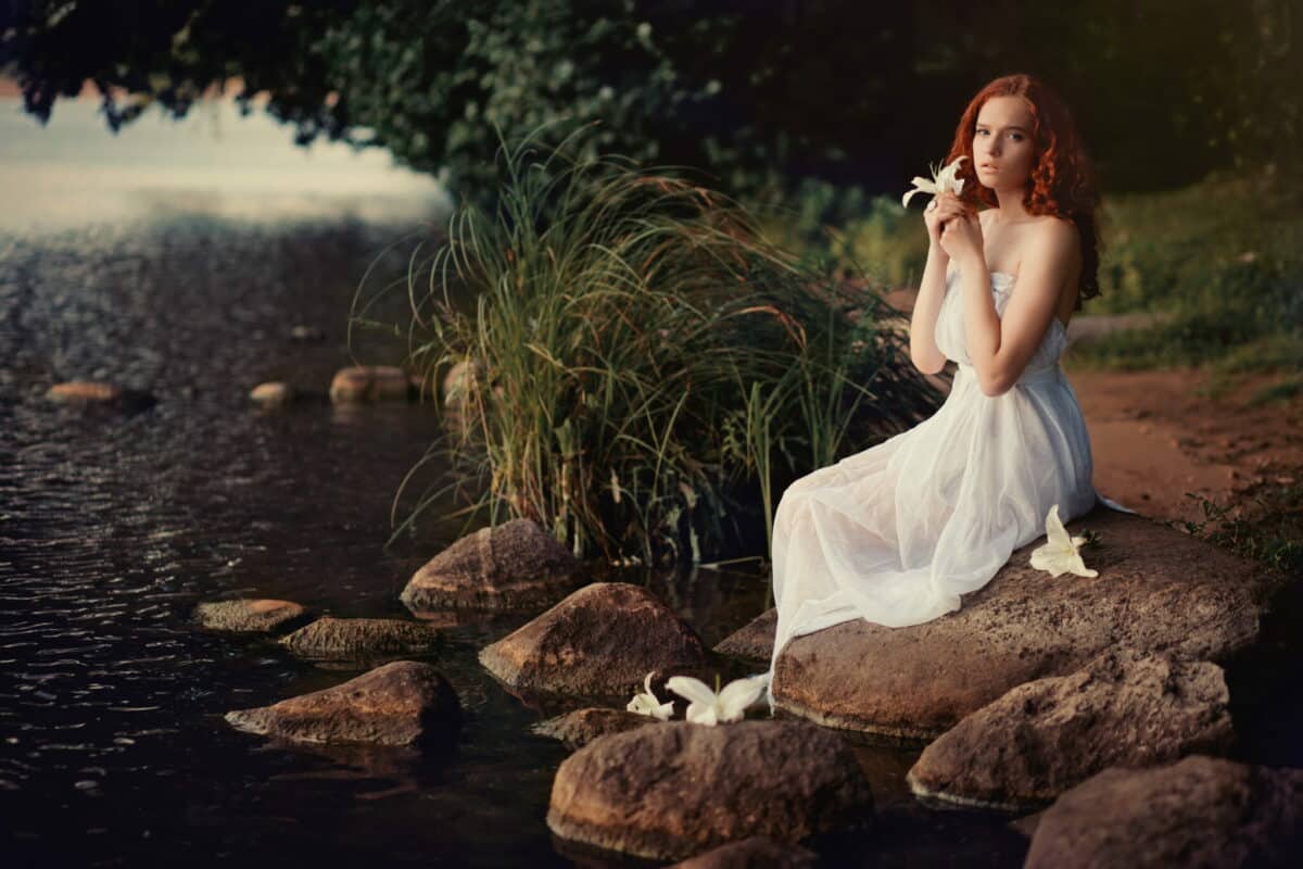 beautiful redhead nymph sitting on a rock by the river, holding a white lily
