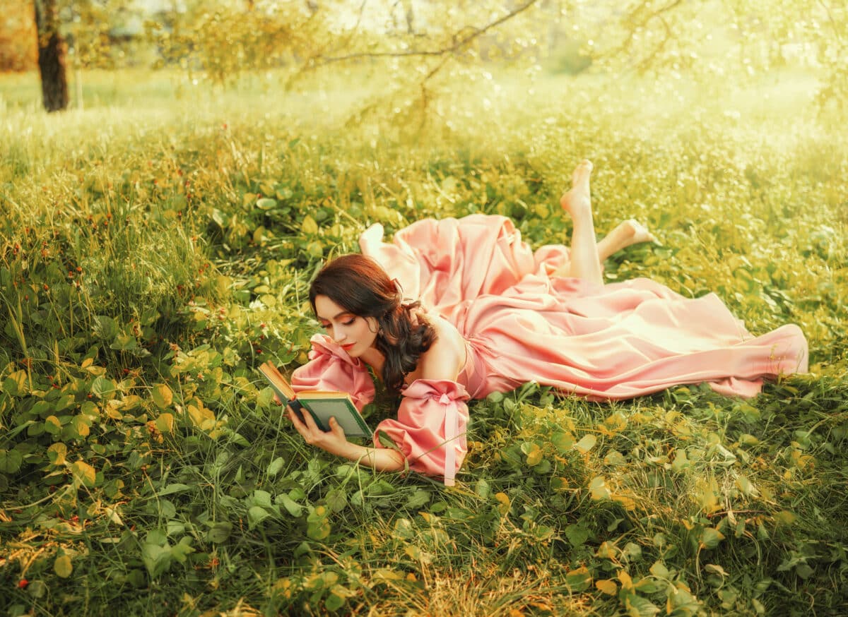 Fantasy happy woman lies on green grass of forest lawn holds in hands magic book novel old style, bright magical sun light. Romantic cute sexy girl in long pink vintage dress. Medieval lady magician