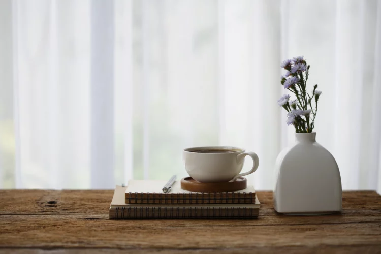 white coffee cup with diary notebook and flower in vase on wooden desk.