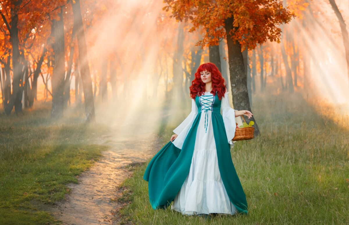 bright creative photo of young beauty, girl in white dress with green cape in bright bottom forest with basket of juicy ripe apples, fabulous princess with closed eyes and red healthy wavy hair