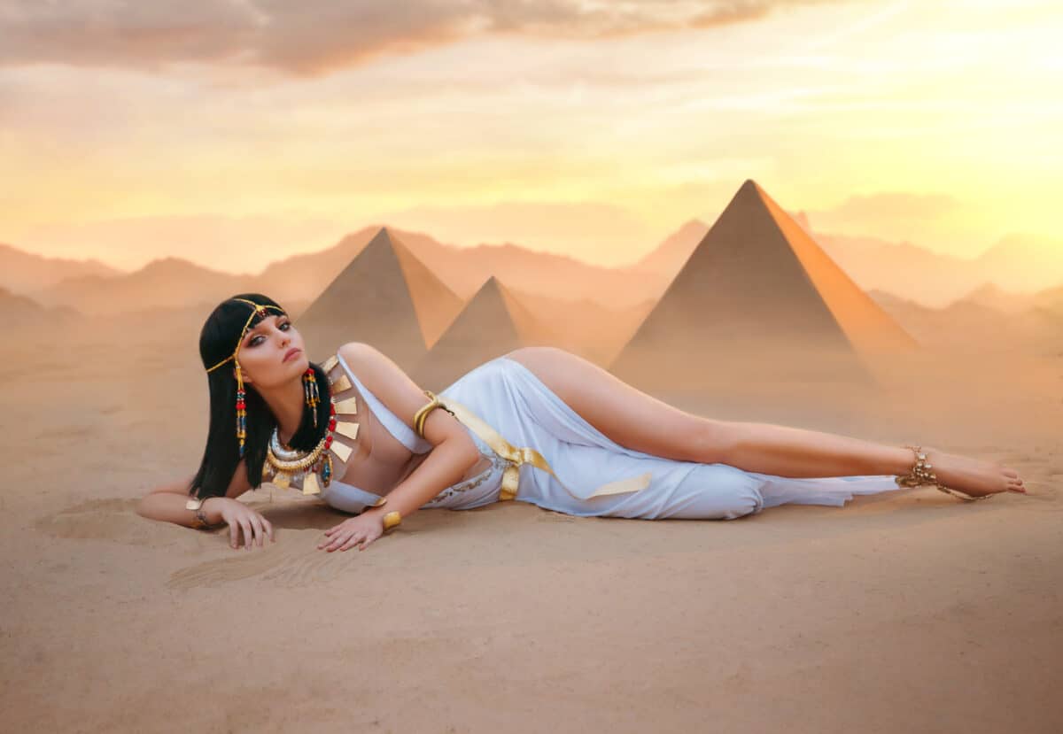 Egypt Style Rich Luxury Woman. Sexy beautiful girl goddess Queen Cleopatra lies on yellow sand desert pyramids. Art ancient pharaoh costume white dress gold accessories. Black hair wig Egyptian makeup