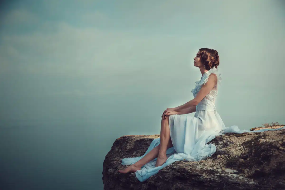 Beautiful bride sitting on the edge of a cliff on the coast with angelic dress