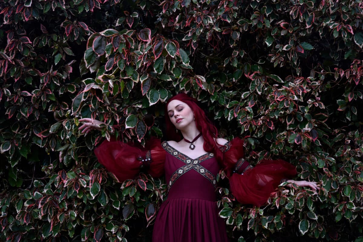 portrait of pretty female model with red hair wearing glamorous renaissance red ballgown. Posing in a fairytale castle location with staircases