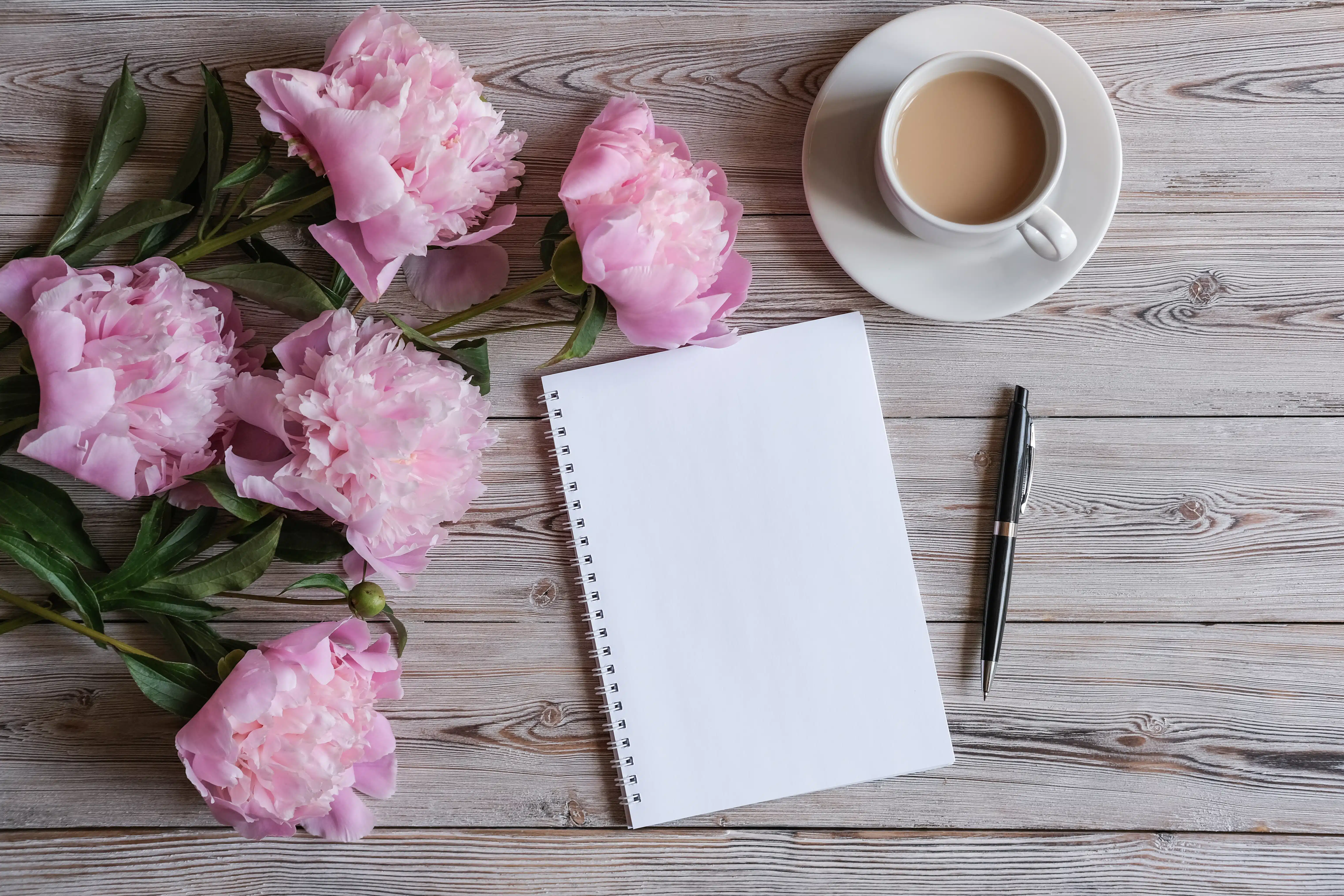 Top view of feminine workspace with blank notepad, cup of coffee and peonies flowers. 