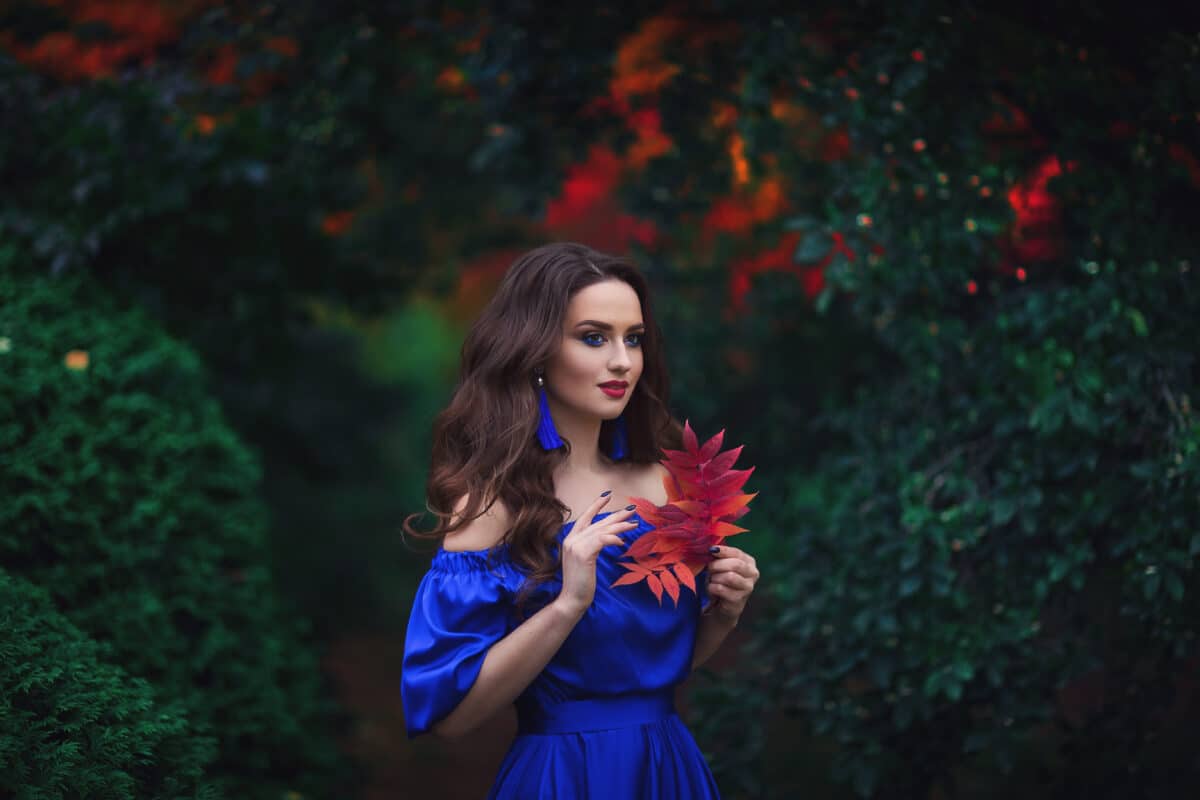 Romantic portrait of a beautiful young girl with blue makeup and red lips, in a long ultramarine blue dress with long blue earrings, on the background of an autumn рark with red leaves