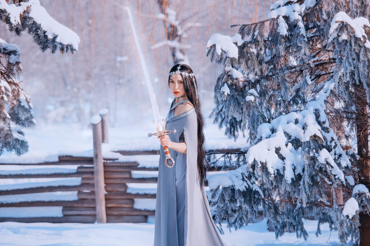 protector on alert, fight of good and evil, magnificent courageous girl with dark long hair in gray warm cloak with glowing crystal sword, brave war goddess in cold winter white forest, snow queen