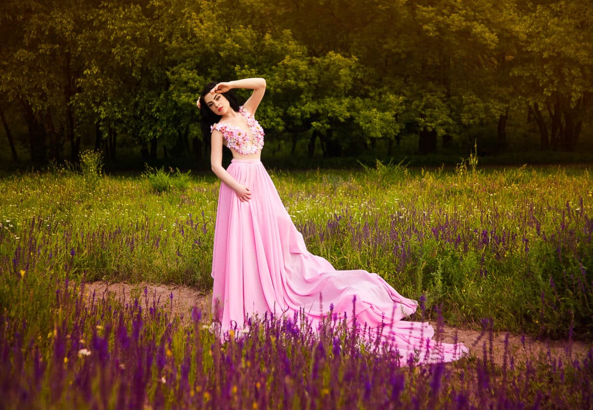 Beautiful brunette woman with long hair in a pink chic flowering dress stay on green grass with purple field flowers in the field with closed eyes. Fantasy portrait.