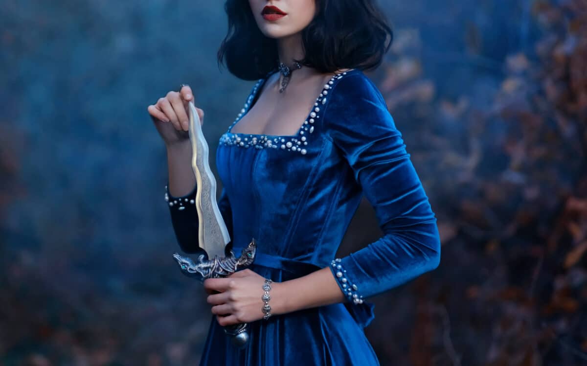 face cropped, red lips close-up. Fantasy medieval woman warrior queen holding dagger, knife in hands. velvet vintage blue dress. Girl princess vampire, brunette short hair. Nature forest dusk night