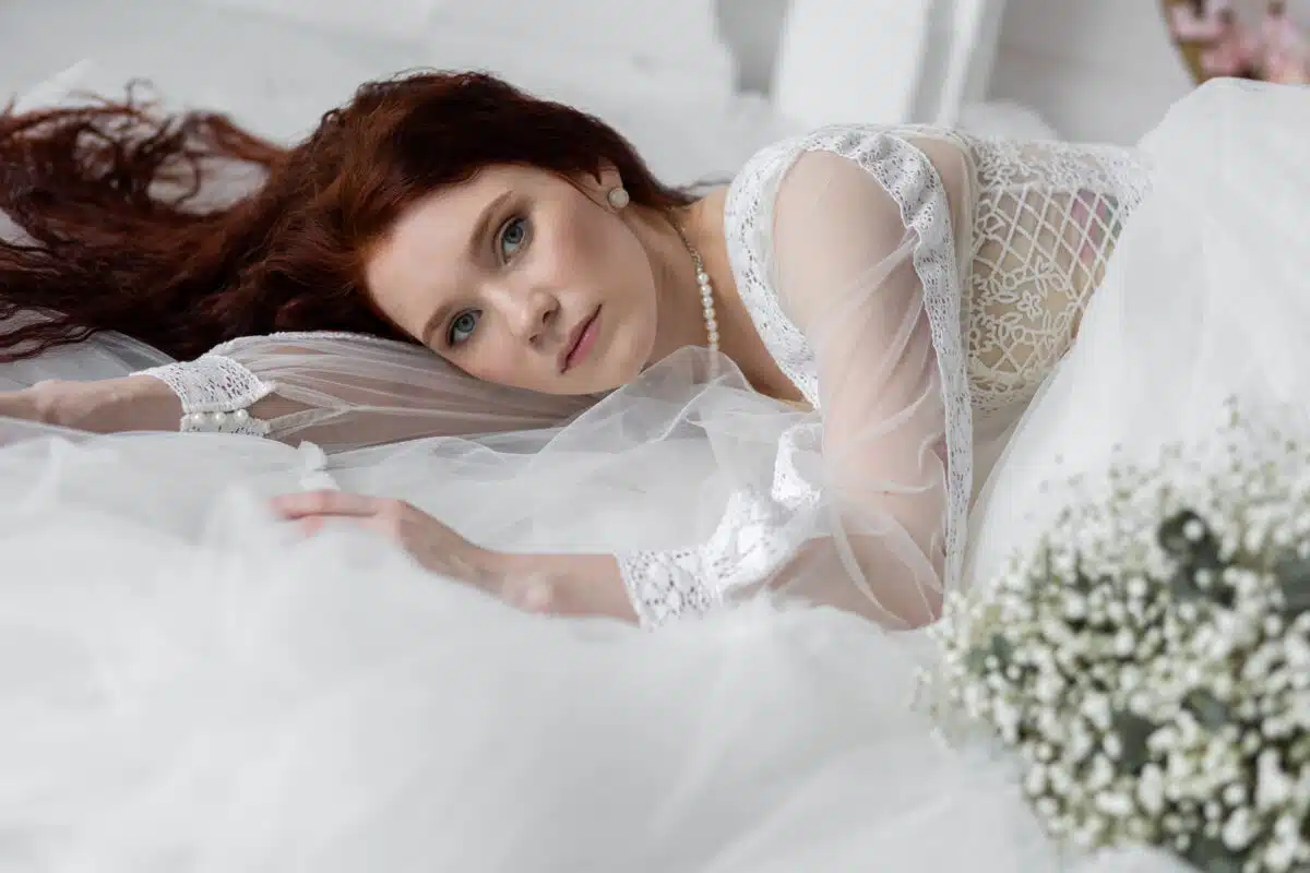 a beautiful redhead with a long hair in a white lace dress and pearls lying on the bed