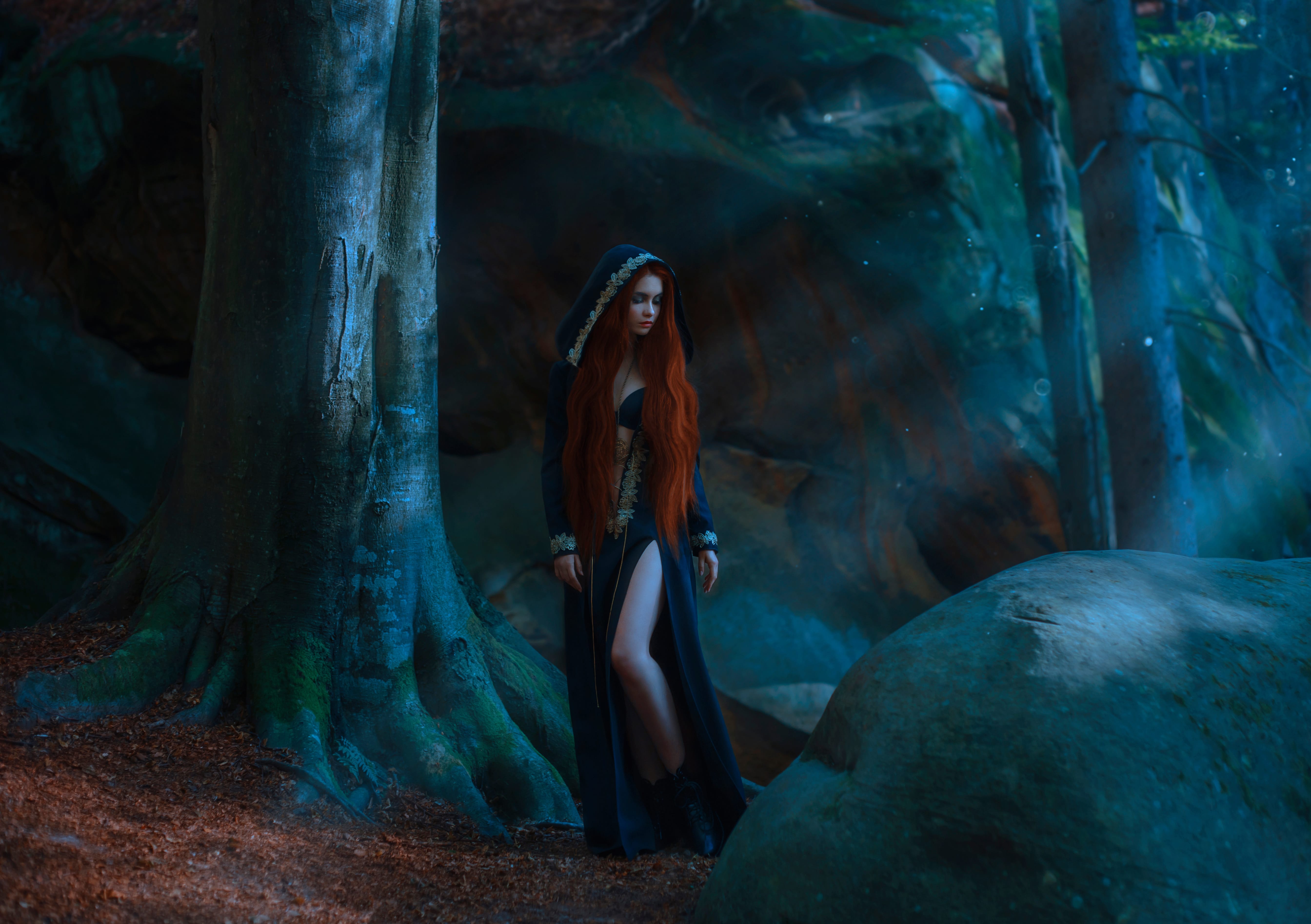 Red-haired attractive slender girl in a long dark blue dress, cloak with a hood and open chest and leather boots standing in a dark forest in the rays of moonlight. the young princess has gone astray