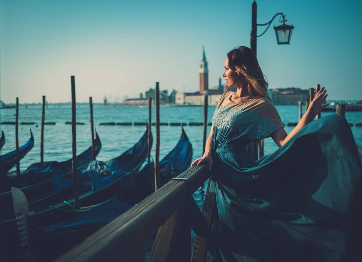a lady in blue green dress is standing by the sea with gondolas