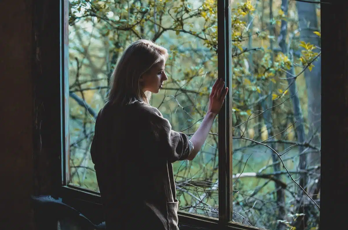 Pensive woman standing by the window, woods in view outside