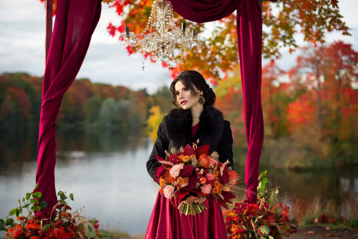 A beautiful bride in a dark red dress stands under a large tree with red and yellow leaves. A destination autumn wedding, romantic country ceremony with marsala decor.