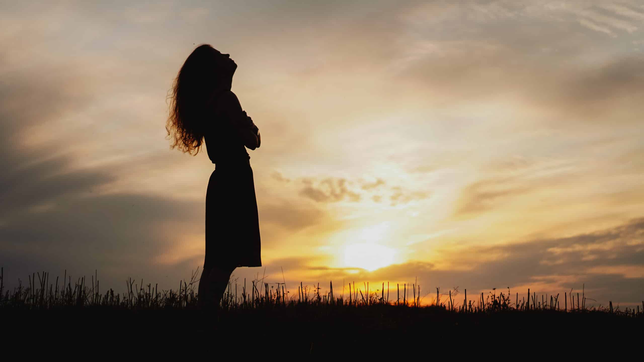 Silhouette of a young woman standing in dry grass field on sunset