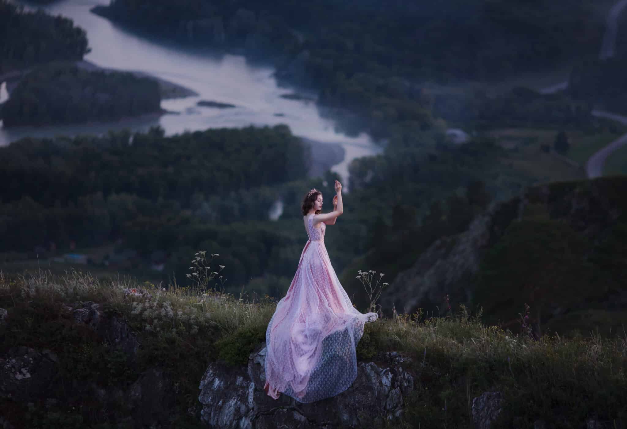 A girl in a pink dress with a long train is dancing on the top of the mountain