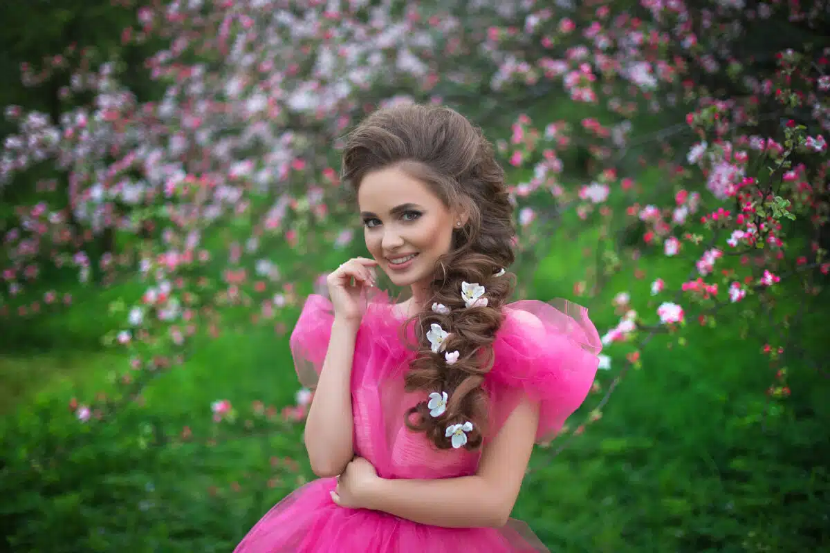 A beautiful young girl with long hair in a light pink ball gown walks through a blooming apple orchard