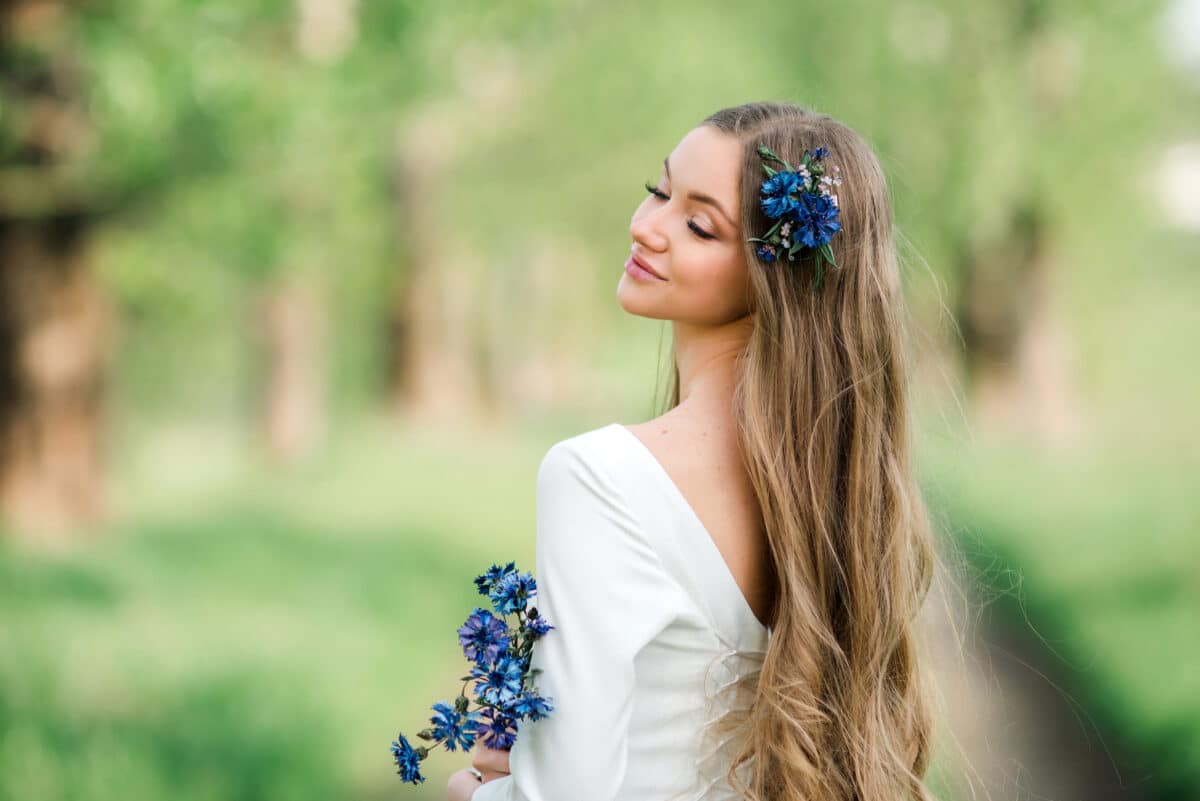 A young happy girl with long hair in a white dress with cornflowers enjoys the summer nature