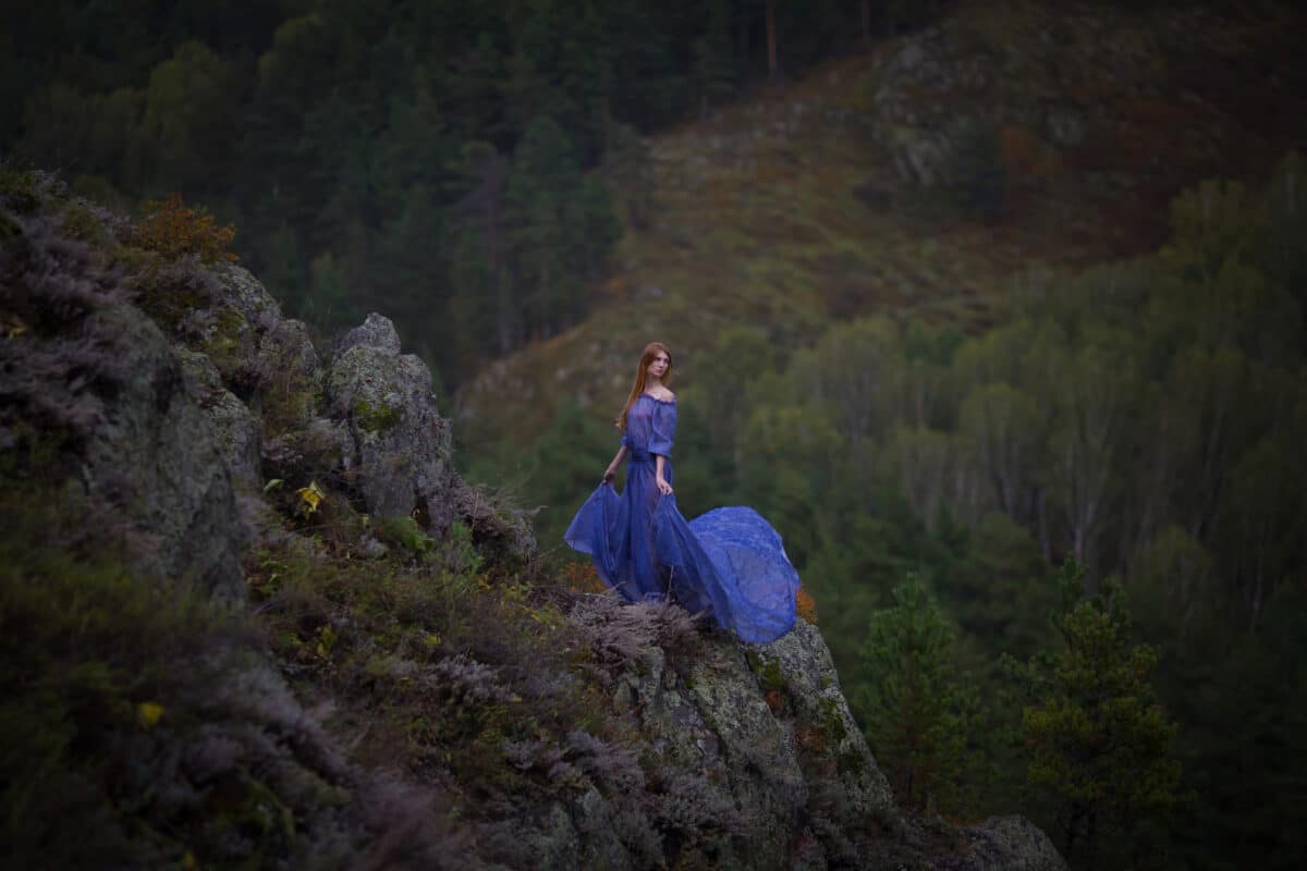 A beautiful red-haired girl in a blue dress walks in the mountains. Portrait.