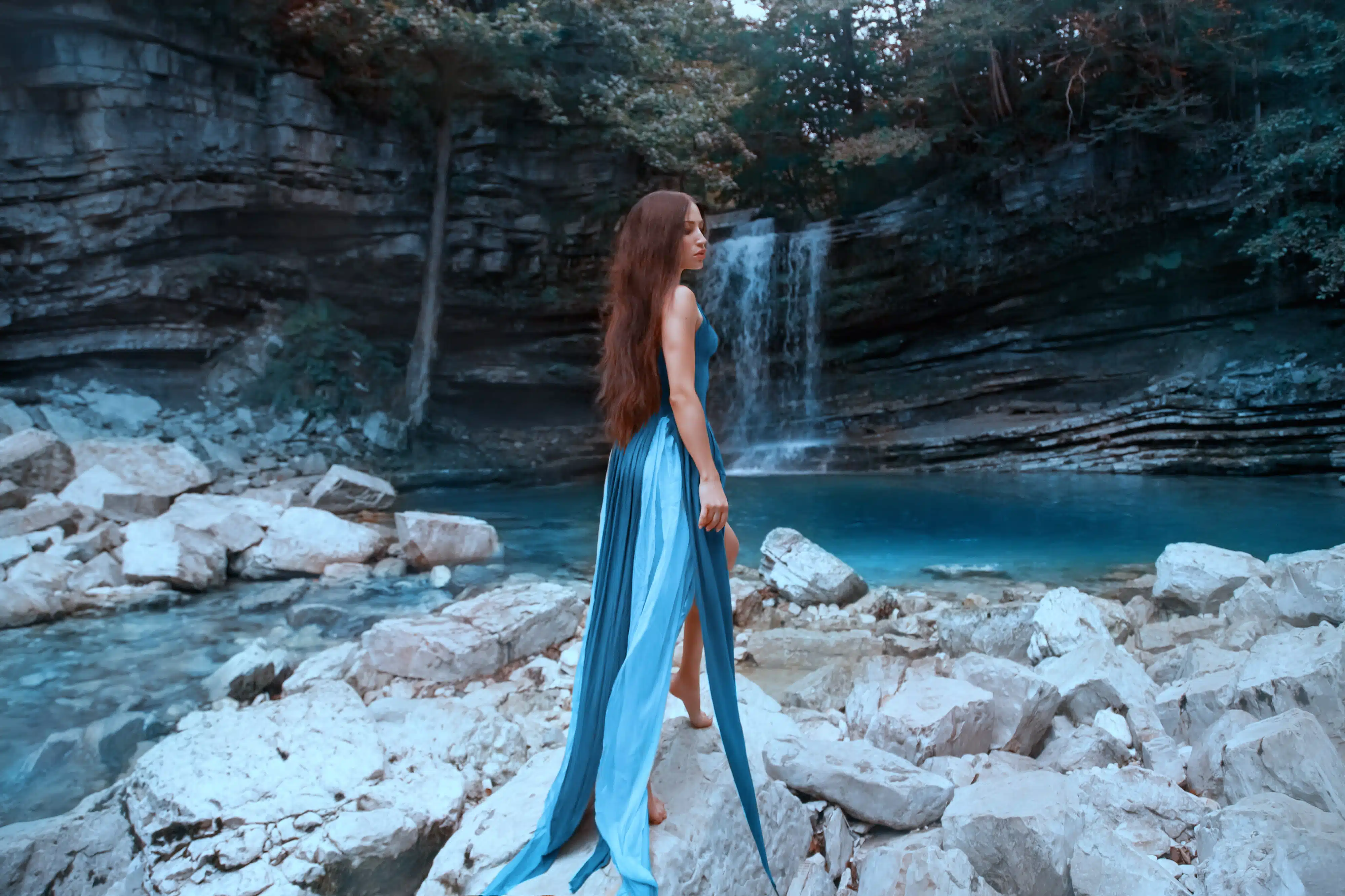 charming girl with long luxurious healthy black hair posing in front of high waterfall in lagoon with blue clear water in Martvili Canyon, lady in light flying dress of heavenly color with cut train