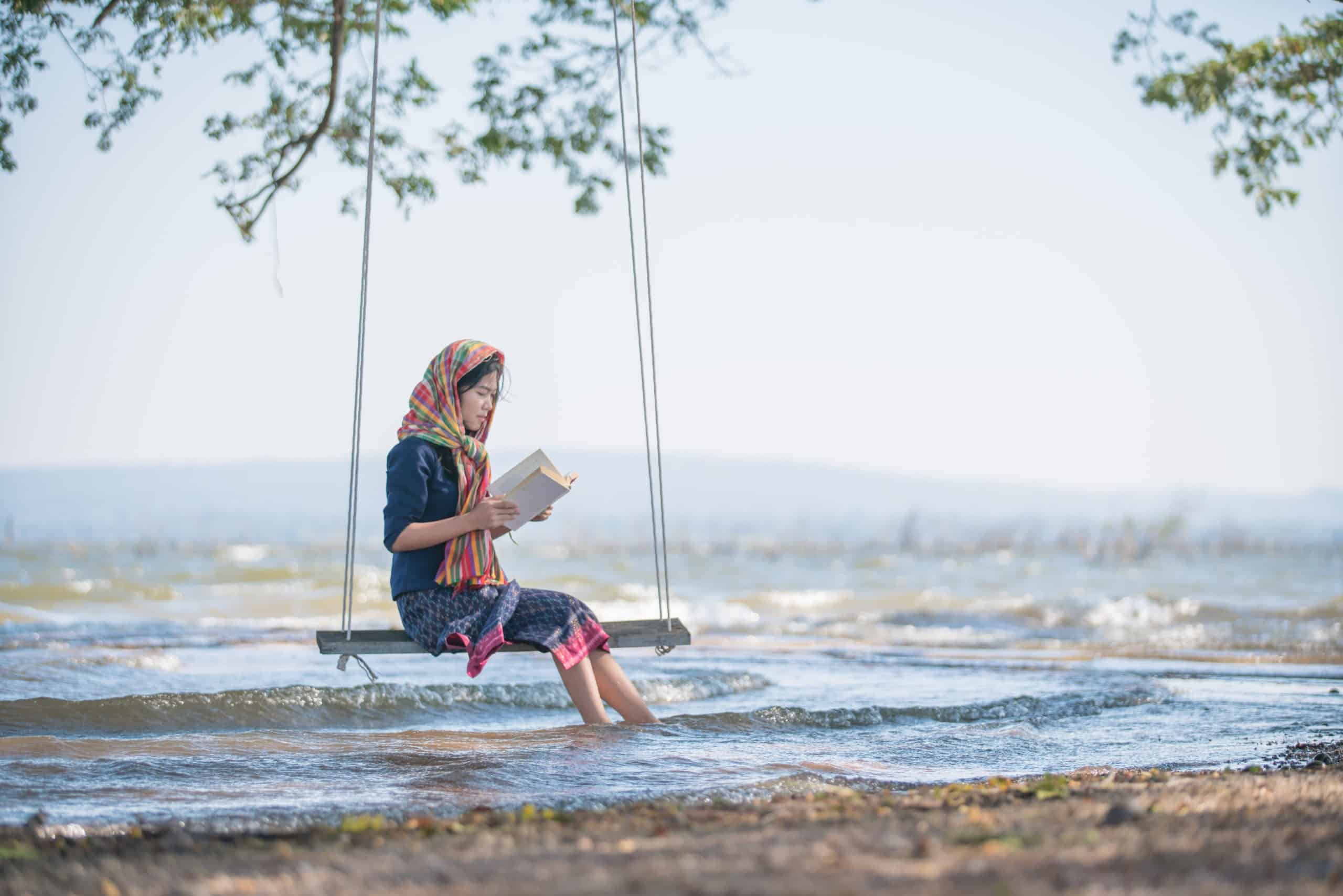Asian woman in traditional dress reading while sitting on a swing by the sea.