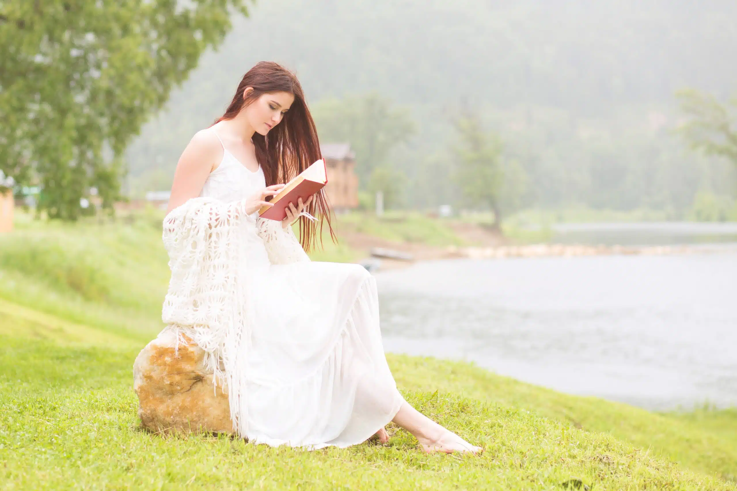 Woman in white dress reading a book by the lake.