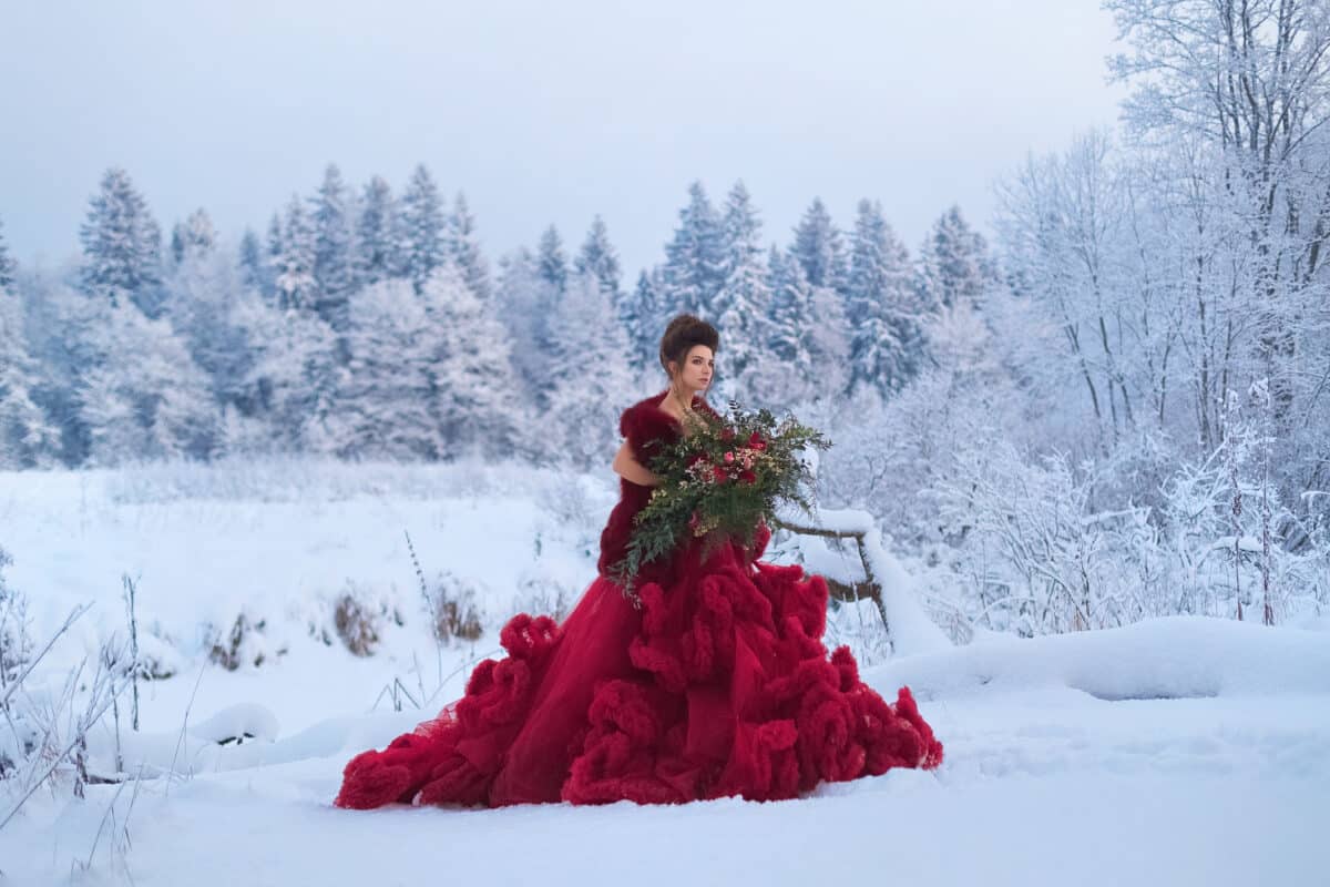 A young royalty in a lush Marsala dress with a large bouquet of red flowers in the snow