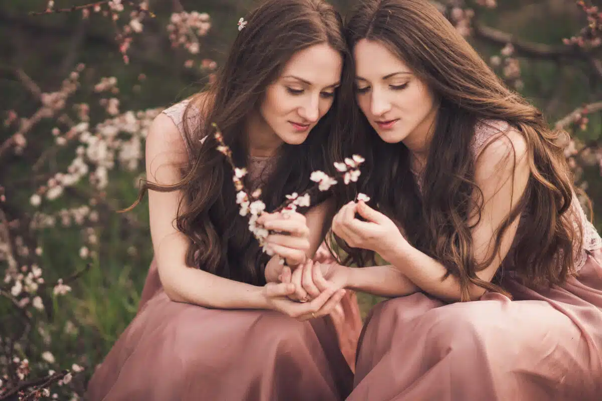 Beautiful happy young twins sisters in long evening dresses enjoying smell in a blooming spring garden. Having fun together, positive emotions, bright colors. Copy space.