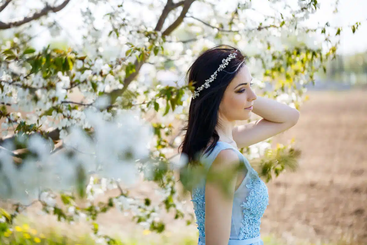 a beautiful girl in a light summer blue long dress adorned in her hair against a blossoming tree