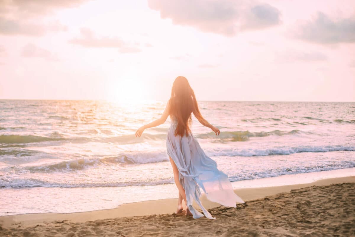 mysterious mermaid with long black hair slowly walks in water of ocean, sea nymph listens to wind like wave in long blue dress with flying train, looks at divine sunset.