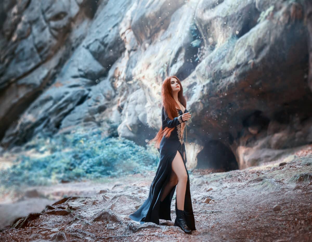 red-haired cute girl in long black dress with bare legs, open belly and golden weaving walks among icy cold rocks on ground with tree roots, girl with her eyes closed in bright glare of sun