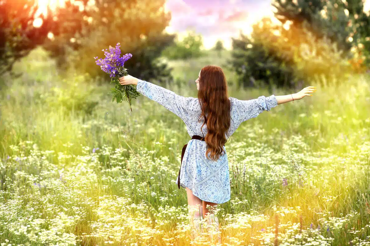 A young redhead lady standing in the field of wildflowers holding a bouquet of purple flowers, arms raised and extended to either sides