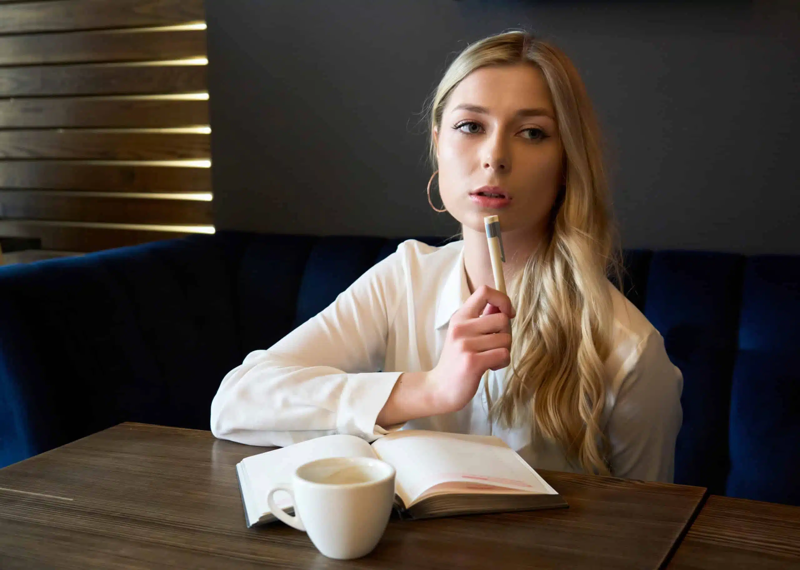 pretty blonde woman thinking, writing in notebook, mug with drink on the table.