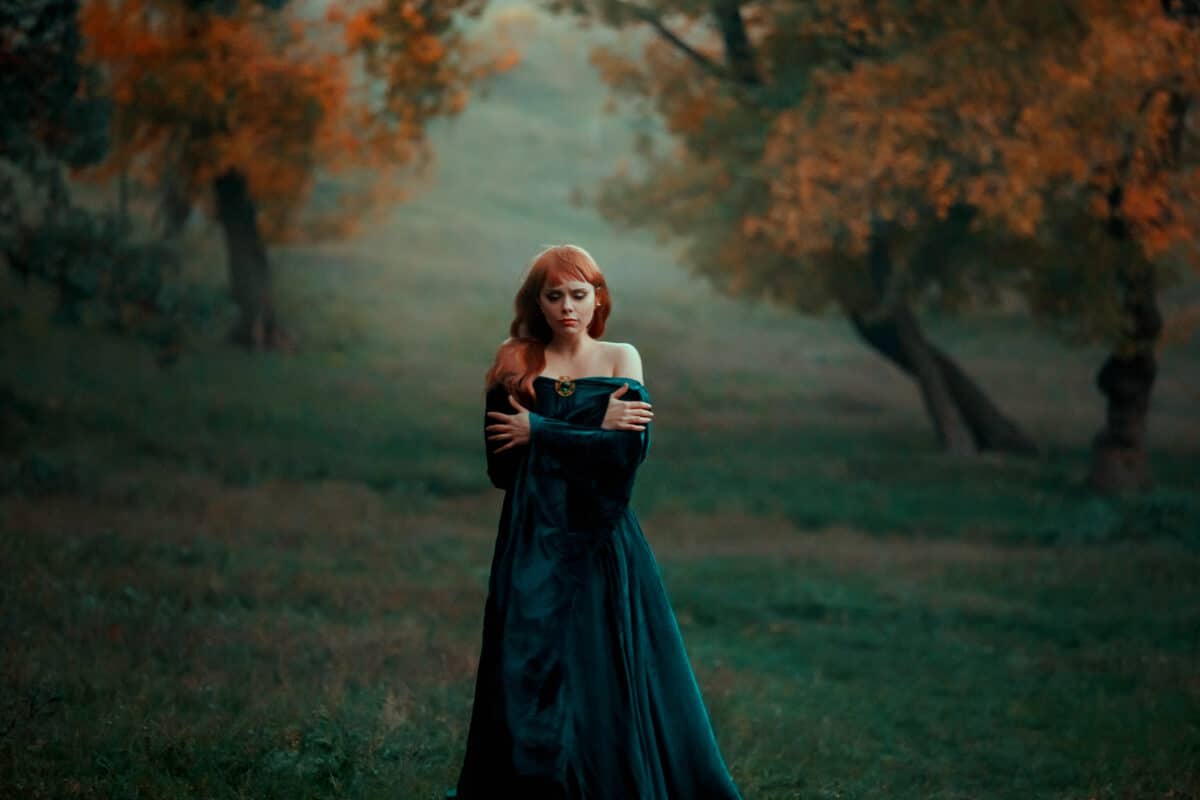 lonely sad girl walks alone in terrible dark dangerous forest in long green emerald dress and raincoat with open shoulders, princess got lost and froze, young witch with red hair looks for path home