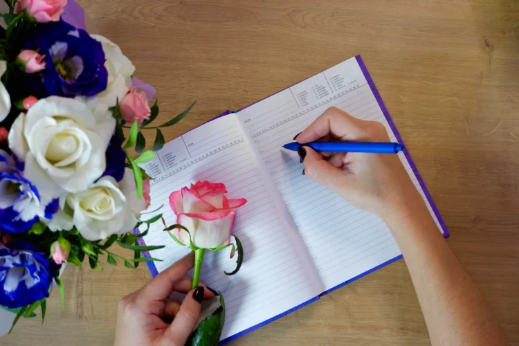 Female hands writing in open notebook and bouquet of roses on old wooden table. 