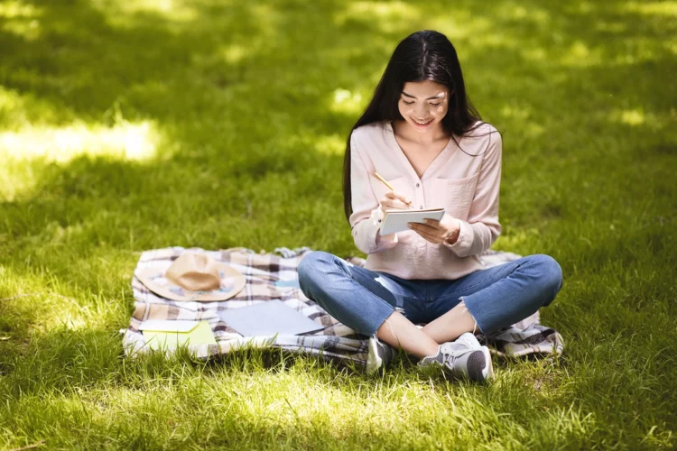 Smiling asian girl making notes in notepad while sitting outdoors in park.