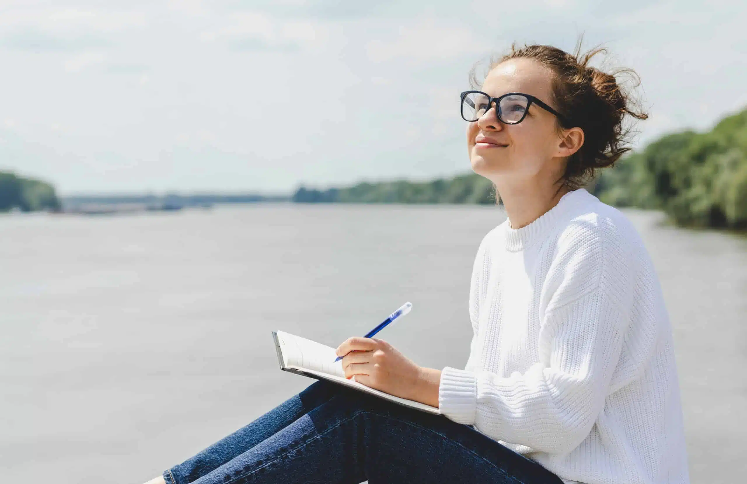 Young woman in glasses writes in notebook outdoors by the lake.