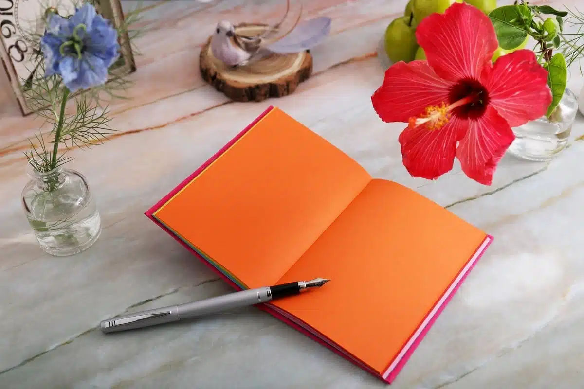 Orange blank page notebook, pen, and hibiscus flower on rustic table setting.
