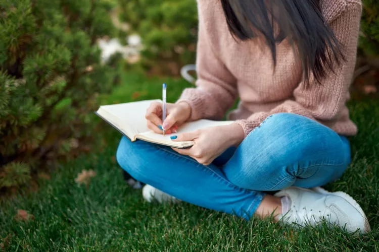 long-haired female writing in a notebook in lotus position on the grass.