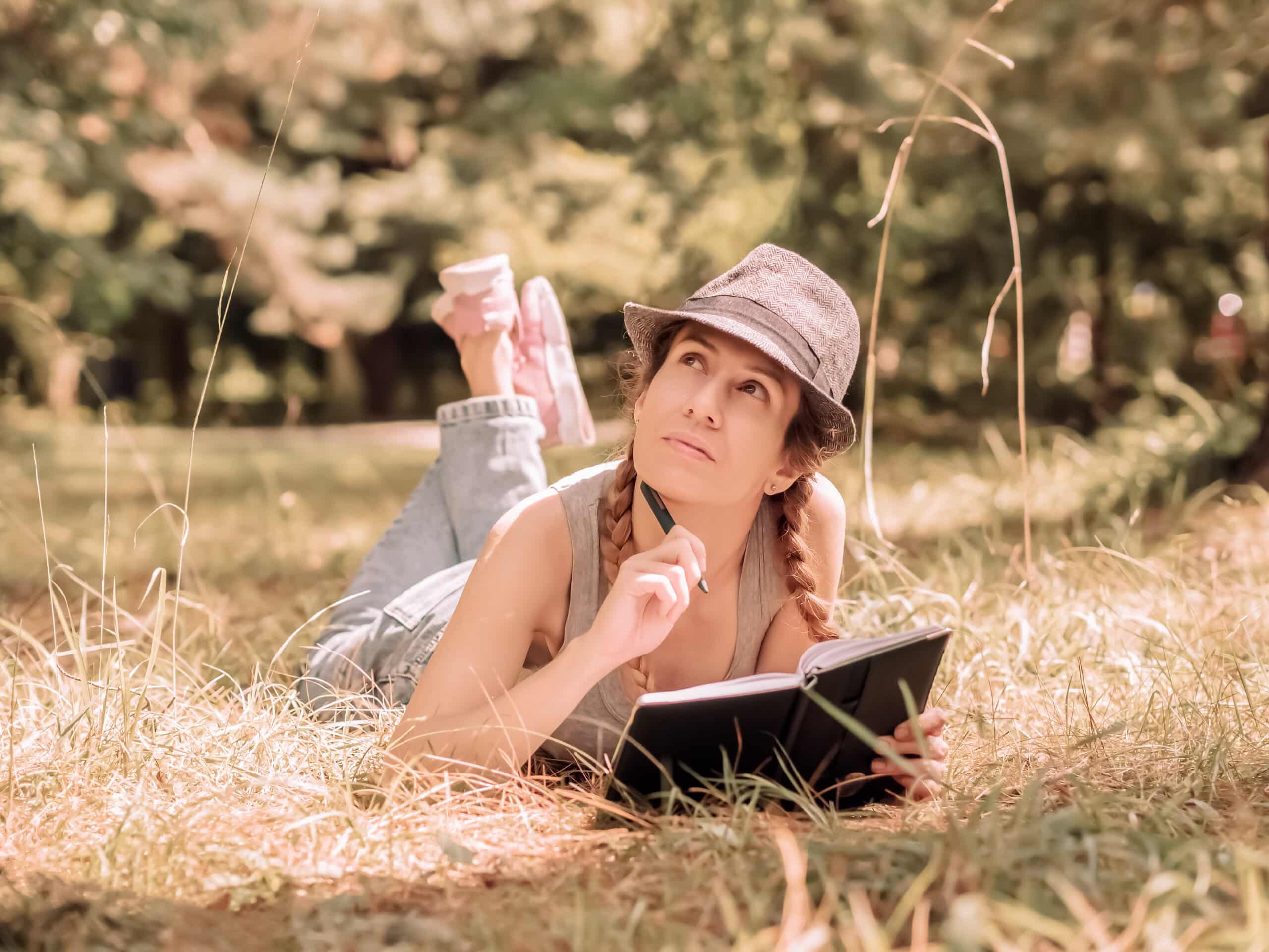 A young woman in a hat lies in the grass in a sunny forest and looks up thoughtfully, holding a pen and a notebook in her hands.