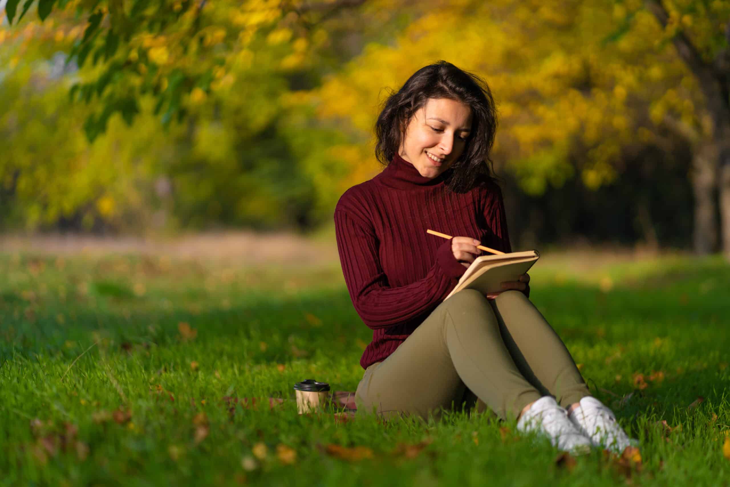A nice woman writes in her notebook, sitting on a lawn in an autumn park