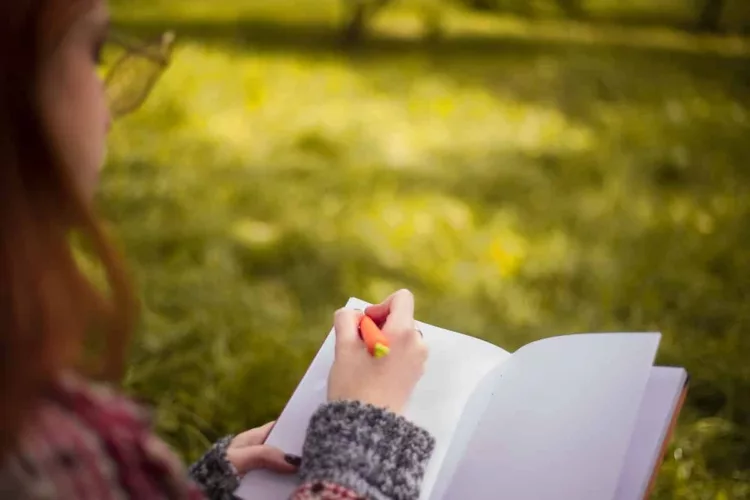 cute redhead reading a book and writing outdoors in park under big tree on grass.