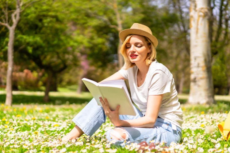 A young blonde in a hat reading a book in spring in a park in the city