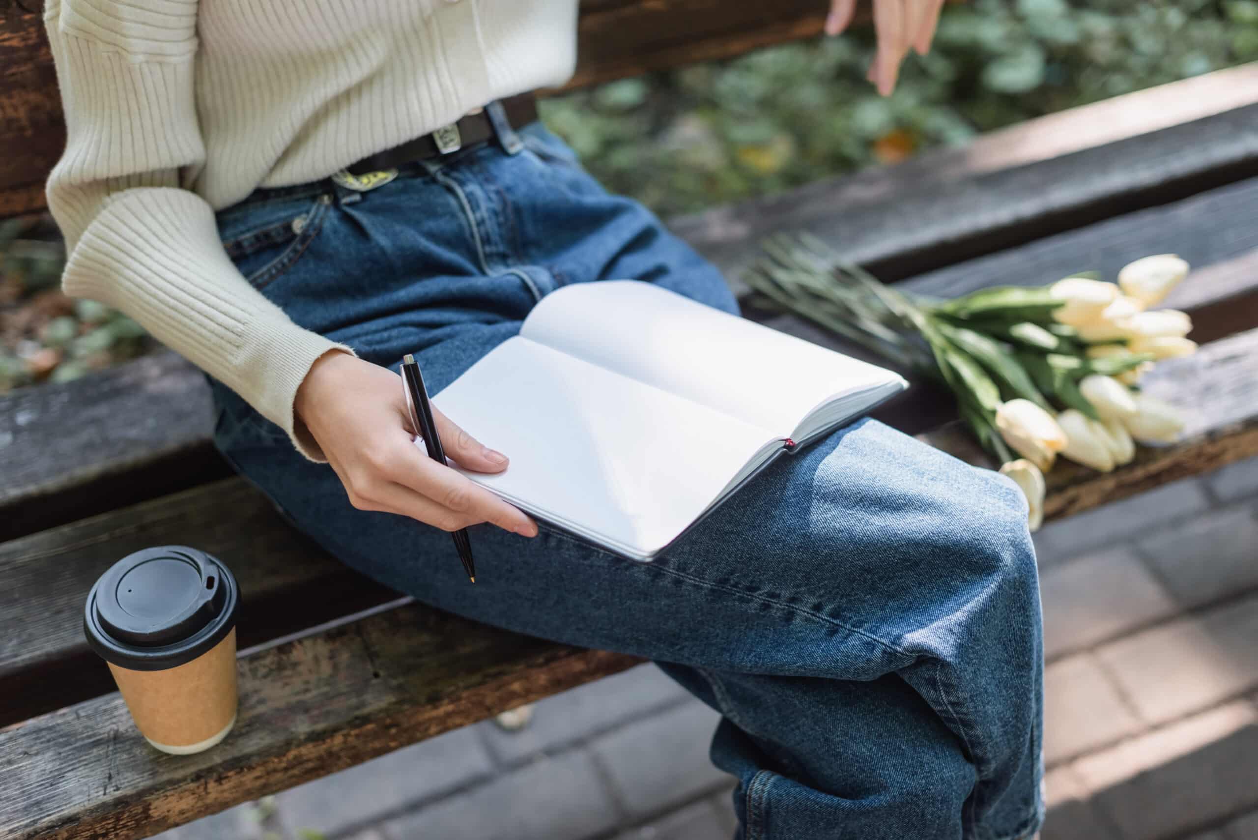 woman in jeans holding notebook near bouquet of tulips and paper cup on bench.
